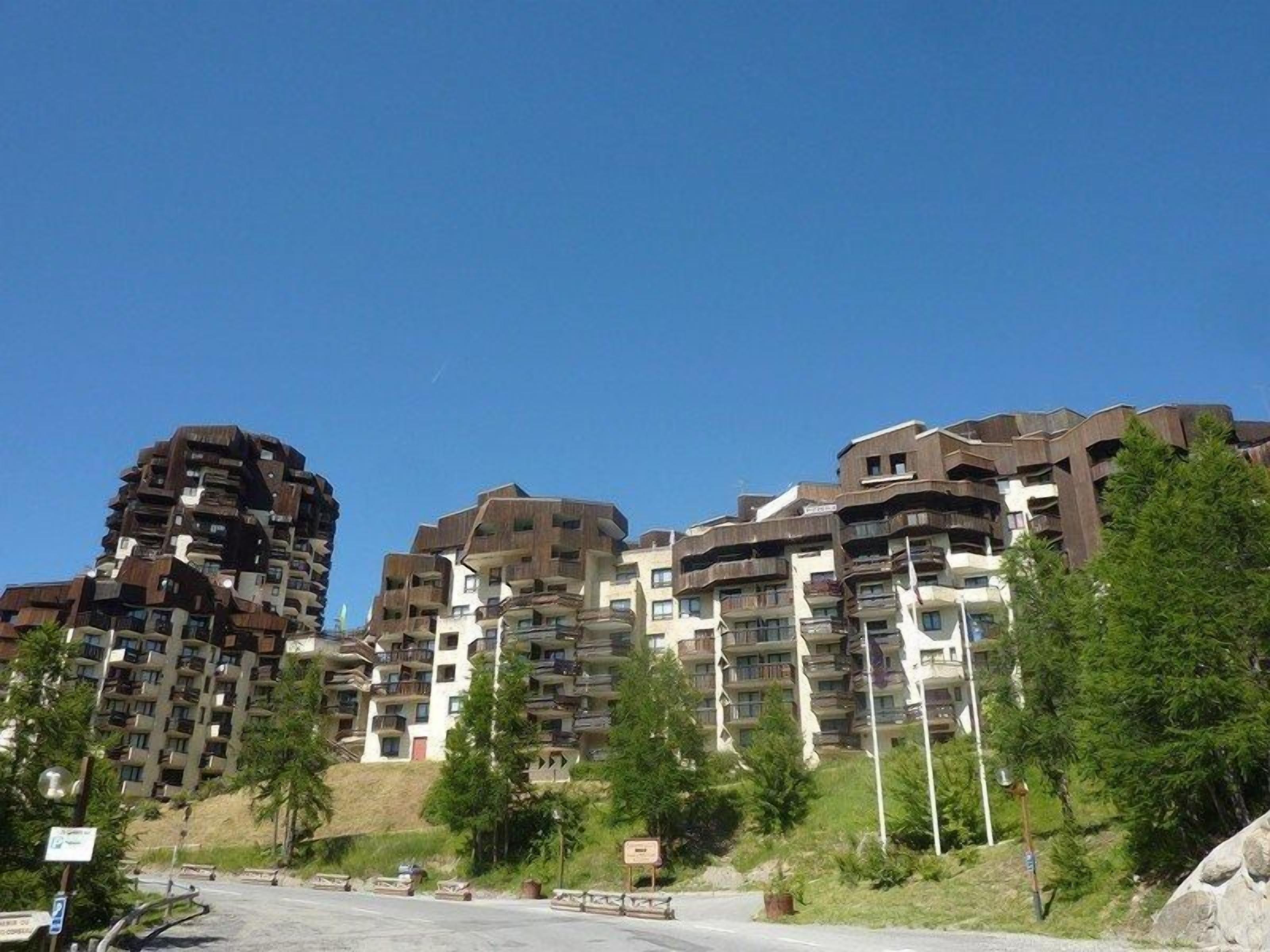 A residential building with multiple balconies, a clear sky, and some greenery in the foreground.