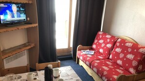 Ett mysigt rum med en röd soffa, ett träbord med muggar och snacks, en tv på väggen och ett fönster med gardiner.
