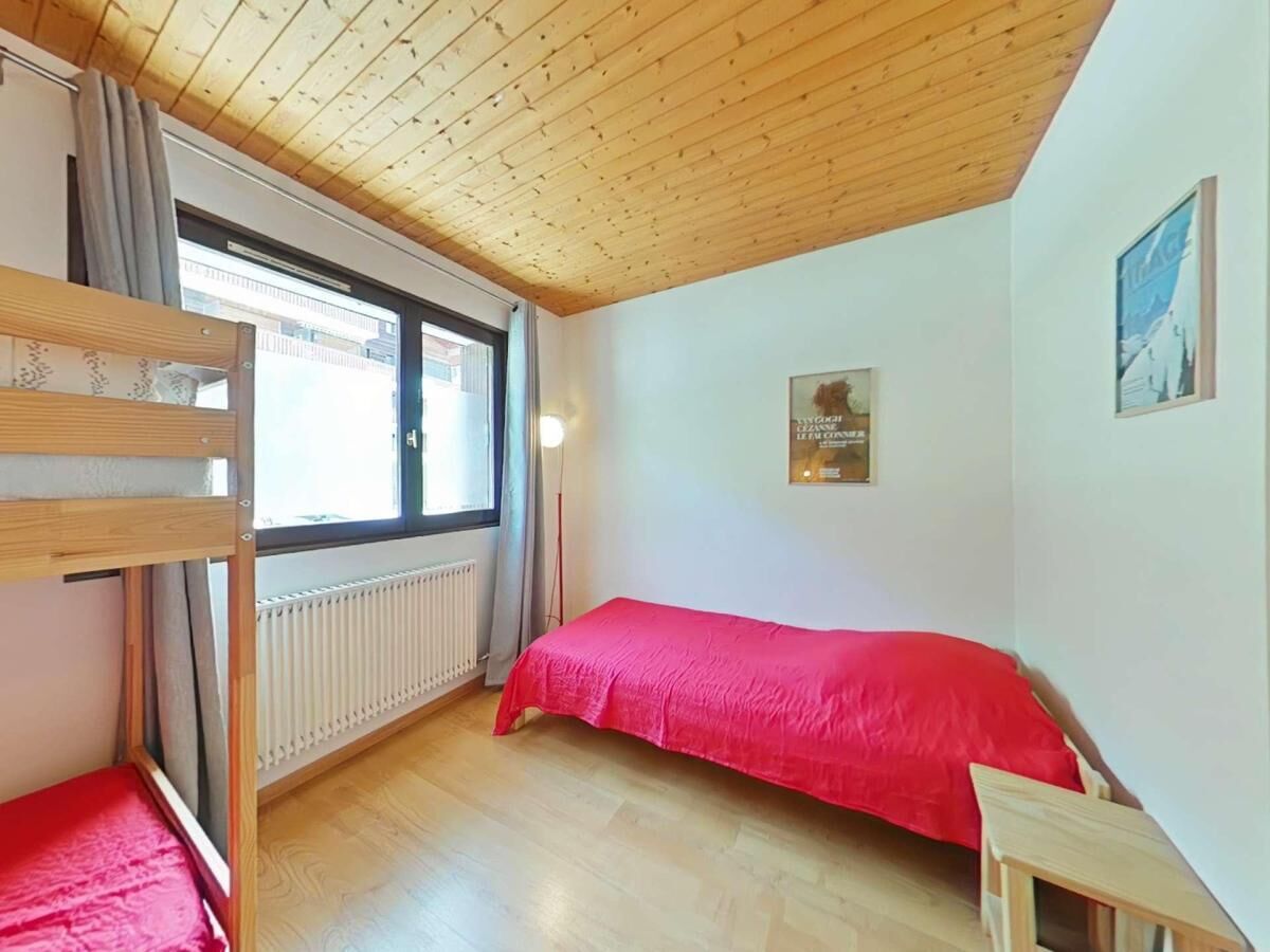 A room with a bunk bed, a single bed with a red blanket, a radiator, a window with curtains, and a framed picture on the wall.