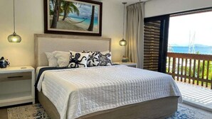 Une chambre avec un grand lit, un ventilateur de plafond, un tableau représentant une plage et une vue sur l’océan depuis un balcon.