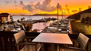 Une vue d’une marina avec des bateaux amarrés, une table et des chaises au premier plan, et un coucher de soleil en arrière-plan.