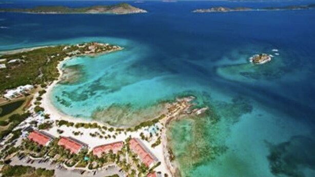 Vue aérienne d’une station balnéaire avec des eaux turquoise cristallines, une plage de sable et une petite île abritant des bâtiments.
