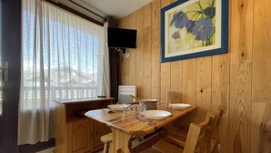 Een houten eettafel met stoelen, een aan de muur bevestigde televisie en een raam met gordijnen.