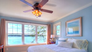 Un dormitorio con una cama grande, un ventilador de techo, una ventana con vista al agua y un cuadro en la pared.