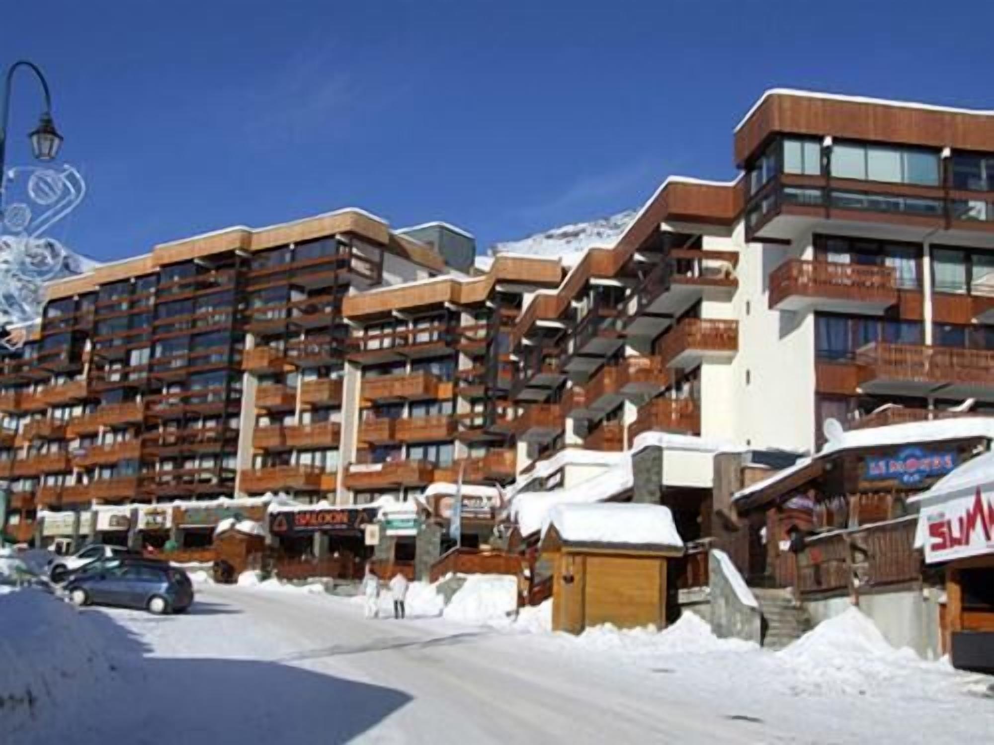 Een hotel met meerdere verdiepingen en balkons, een wintersportoord in de verte, en een met sneeuw bedekte straat.