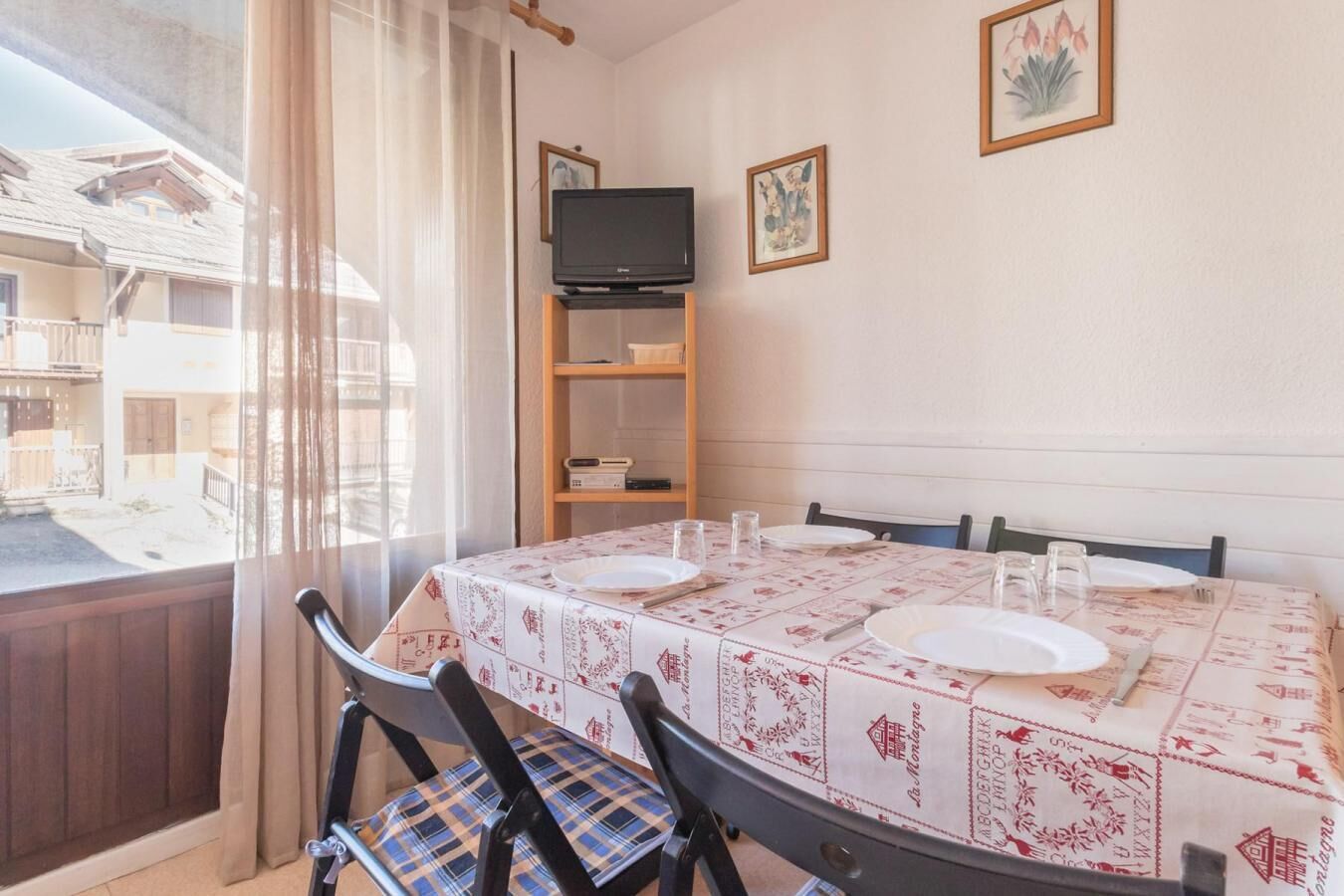 A dining area with a table set for four, a television on a stand, and a window with curtains.