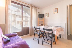 A room with a dining table set for two, a sofa, a TV on a shelf, and a view of a building outside.
