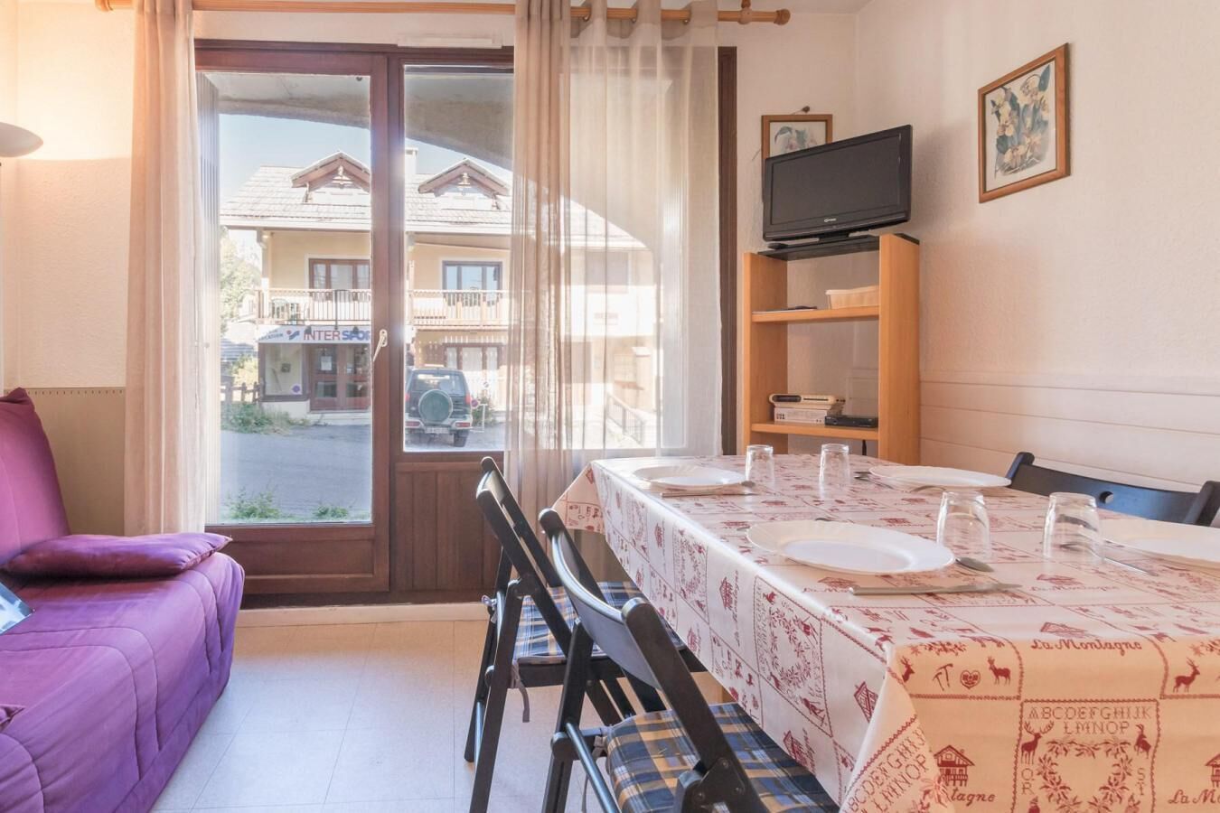 A room with a dining table set for two, a purple sofa, a television on a wooden stand, and a window with a view of buildings and a parked car.