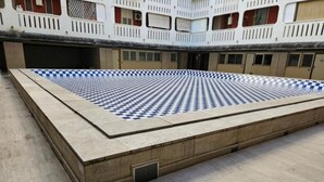 Una piscina con motivo a scacchi, circondata da un muretto e un edificio con balconi sullo sfondo.