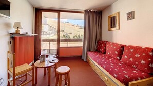 A cozy room with a red sofa, a wooden table, a chair, and a view of a mountainous landscape through a large window. - 50m from ski slopes, 2nd floor, balcony, tv, 38m², Les Menuires (Les Menuires)