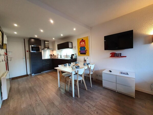 A modern kitchen with a dining area, a flat-screen TV mounted on the wall, and a small white cabinet.