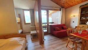 Una habitación con suelo de madera, una cama, un sofá rojo, una mesa de comedor con sillas y vistas a través de un ventanal.