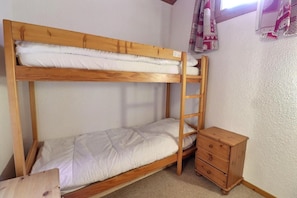 A bunk bed with a wooden ladder, a small wooden dresser, and a window with patterned curtains.
