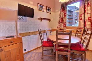 A cozy dining area with a round table and wooden chairs, a television mounted on the wall, and a window with patterned curtains.