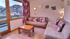 A cozy living room with a sofa, a small table, and a view of a residential area through a large window. - 10m from ski slopes, 3rd floor, balcony, 43m², Méribel-Mottaret (Méribel - Les Allues)