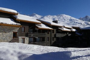 A snow-covered lodge with a stone facade and wooden balconies, set against a backdrop of snow-capped mountains. - 10m from ski slopes, 3rd floor, balcony, 43m², Méribel-Mottaret (Méribel - Les Allues)