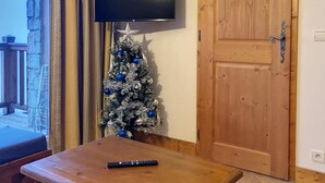 Una habitación con un cofre de madera, un televisor montado en la pared, un árbol de Navidad y una puerta de madera.
