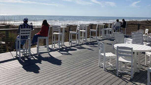 Una terraza de madera con sillas y mesas blancas con vistas a la playa.