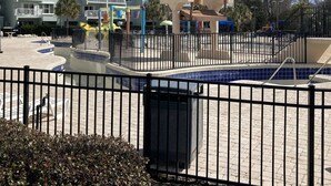 Un edificio de varios pisos con balcones, un área de piscina cercada con un parque infantil, y un cielo despejado.