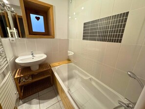 Une salle de bain avec un lavabo blanc, un meuble-lavabo en bois, une douche à parois de verre et un mur carrelé orné d’une fenêtre en forme de cœur.