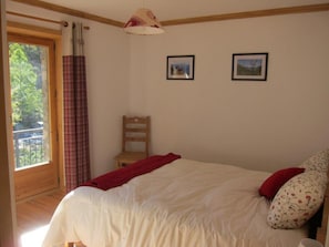 Um quarto com uma cama grande, uma cadeira de madeira, duas molduras com imagens na parede e uma janela com cortinas.
