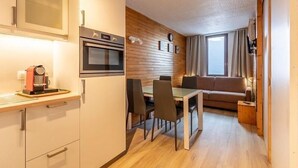 Una cocina compacta equipada con microondas, horno y cafetera. Cuenta con un comedor con mesa y sillas, además de un sofá. La habitación tiene paneles de madera en las paredes y un gran ventanal con cortinas.