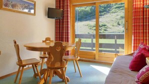 A room with a wooden dining table and chairs, a bed with pink pillows, a television on the wall, and a large window with a view of a green landscape. - 10m from ski slopes, 33m², Méribel-Mottaret (Méribel - Les Allues)