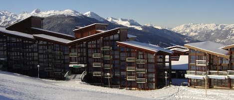 A snow-covered ski resort with wooden buildings, a clear sky, and snow-covered mountains in the background.