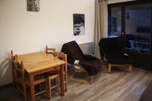 Un coin repas avec une table et des chaises en bois, un fauteuil recouvert de tissu et une vue sur l’extérieur à travers une porte vitrée.