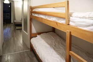 Un lit superposé avec une literie blanche, dans une pièce dotée d’un plancher en bois et d’une porte donnant sur une autre pièce.