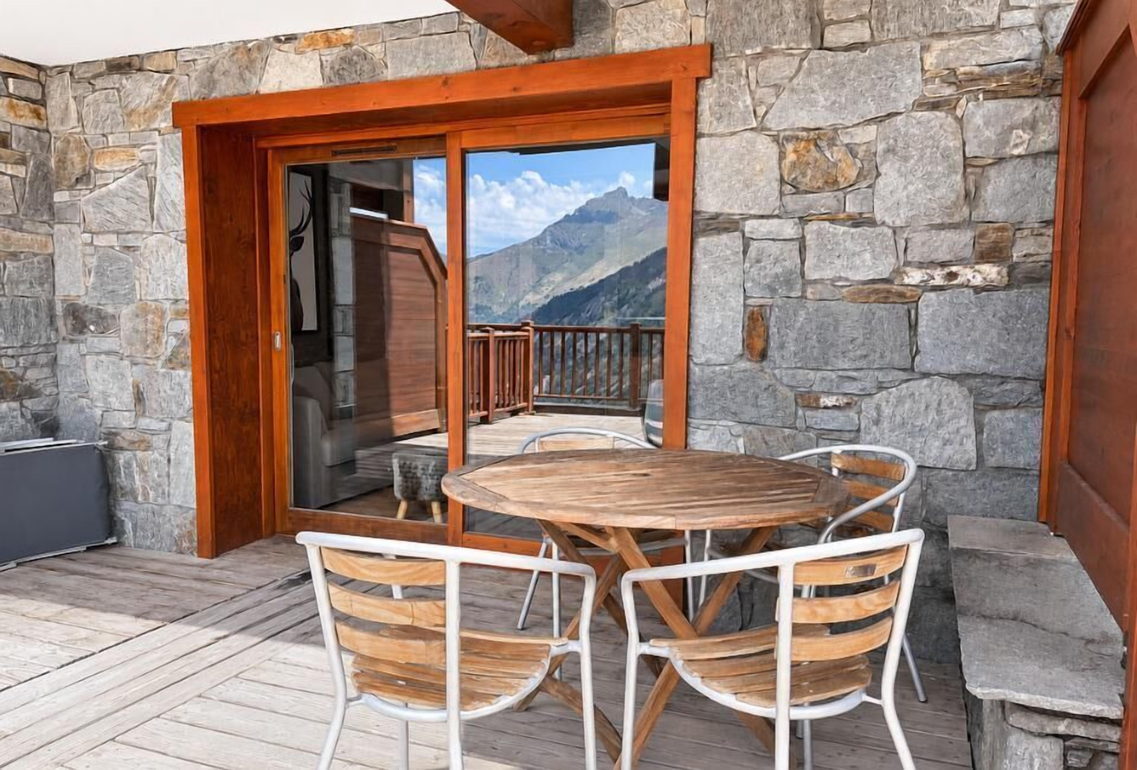 A wooden deck with a stone wall, a round table with chairs, and a view of mountains.