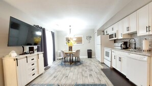 Une cuisine compacte avec des meubles blancs, un téléviseur à écran plat, une table à manger avec des chaises et une petite poubelle.
