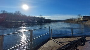 Une terrasse en bois avec une rambarde en métal, donnant sur une rivière, avec des bâtiments et des arbres en arrière-plan.