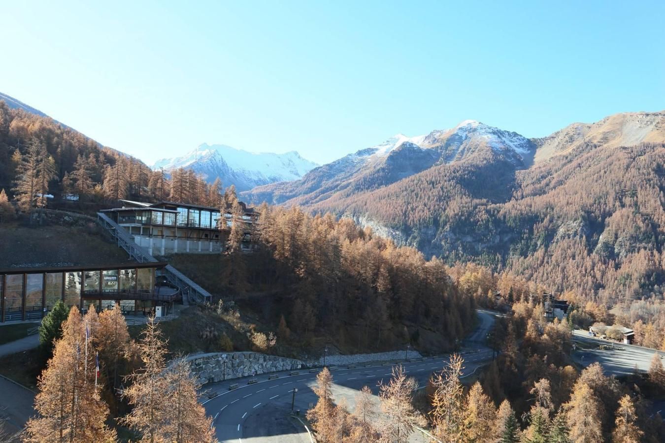 A modern building with a glass facade, surrounded by autumnal trees and mountains in the background.