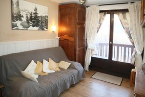 Ein Zimmer mit einer grauen Couch, einem Holzschrank, einem Fenster mit Vorhängen und einem Holzboden.