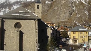 A historic stone building with a bell tower, surrounded by snow-covered mountains and modern buildings.