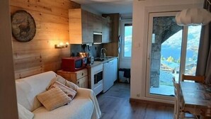 Ein gemütliches Zimmer mit Holzwand, einem weißen Sofa, einer kleinen Küche mit Mikrowelle und Blick auf eine gebirgige Landschaft durch eine Glastür.