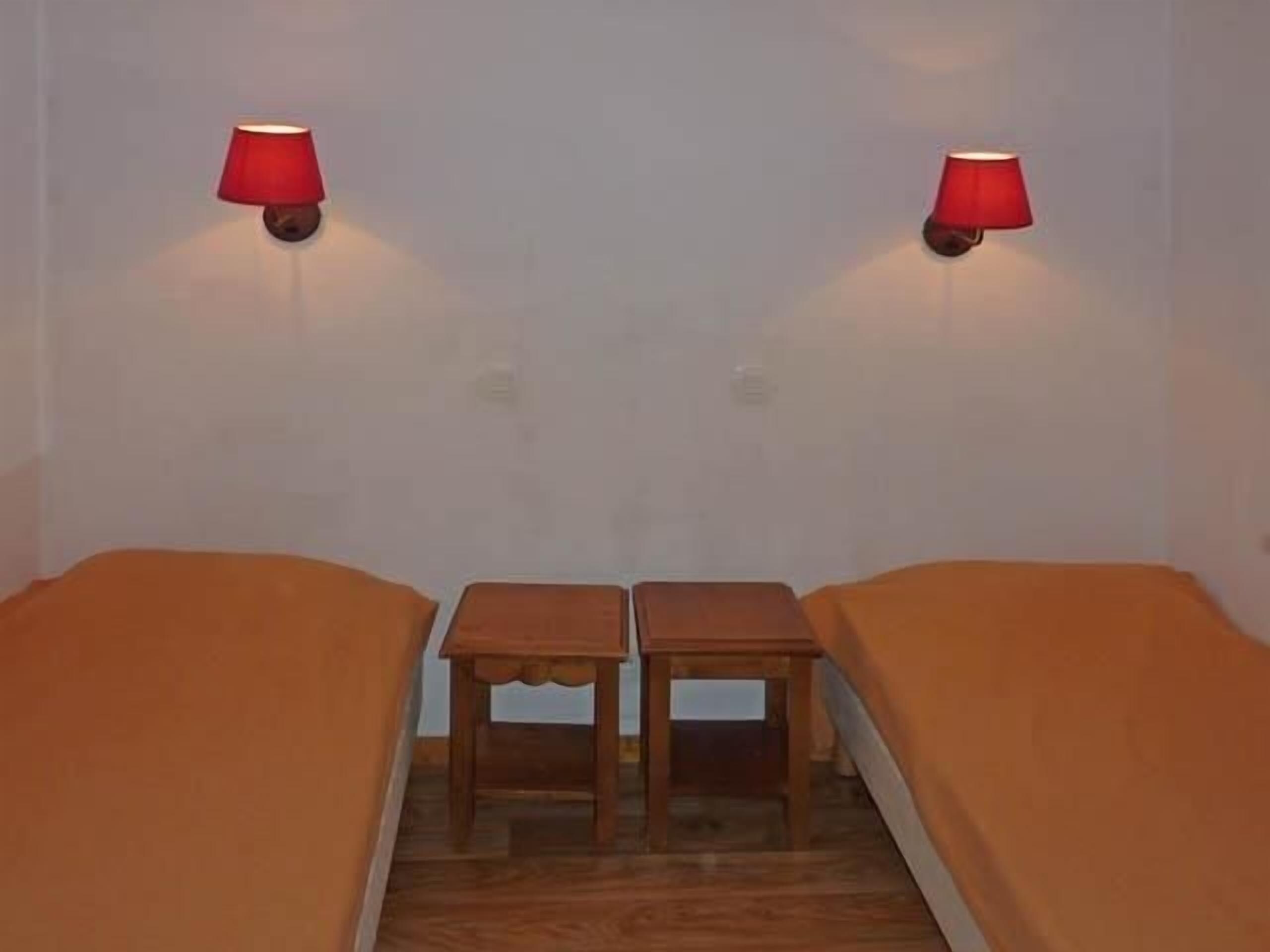 A room with two single beds, a small wooden table, and two wall-mounted lamps.