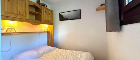 Un dormitorio pequeño con cama, cabecera de madera, estante de pared con figuritas, un cuadro enmarcado y una ventana.