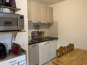 A compact kitchen with a microwave, toaster, and dishwasher. There is a wooden table and chairs, and a shelf with kitchen utensils.