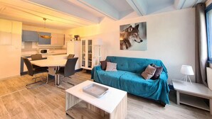 A modern living room with a teal sofa, a dining table with chairs, a kitchen area, and a large wolf painting on the wall.