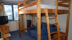 A bunk bed room with a television on a wooden stand, a blue sofa, and a window with a view of buildings.
