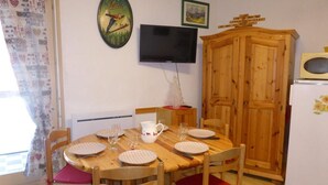 A dining area with a wooden table set for four, surrounded by chairs with red cushions. A television is mounted on the wall above the table.