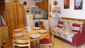 A cozy living room with a wooden dining table set for four, a red sofa with patterned cushions, and a kitchen area with wooden cabinets and a microwave.