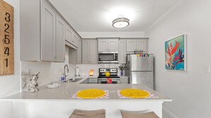 A modern kitchen with white cabinets, a microwave, stove, and a refrigerator. There are two bar stools and a decorative angel statue. - Boho Beach Vibes @ Caribbean Dunes,125 (Destin)