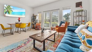 A modern living room with a blue sofa, yellow chairs, a wooden coffee table, and a large TV displaying a beach scene. - Boho Beach Vibes @ Caribbean Dunes,125 (Destin)
