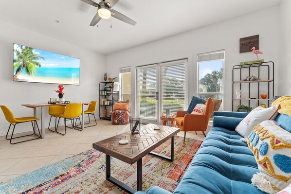 A modern living room with a blue sofa, yellow chairs, a wooden coffee table, and a large TV displaying a beach scene.