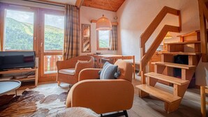 Een gezellige woonkamer met een televisie, rieten meubels, een houten trap en grote ramen met uitzicht op de heuvels.