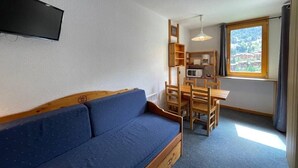 Een compacte kamer met een blauwe bank, een houten eettafel, stoelen, een aan de muur bevestigde televisie en een raam met uitzicht op gebouwen.