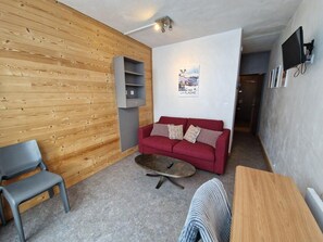 Ein Wohnzimmer mit einem roten Sofa, einem grauen Sessel, einem Holztisch und einem an der Wand befestigten Fernseher.